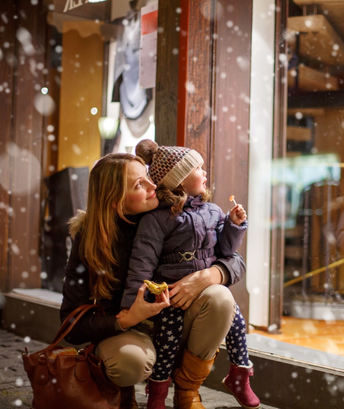 A toddler looks at a festive window while her mother holds her during the holidays.